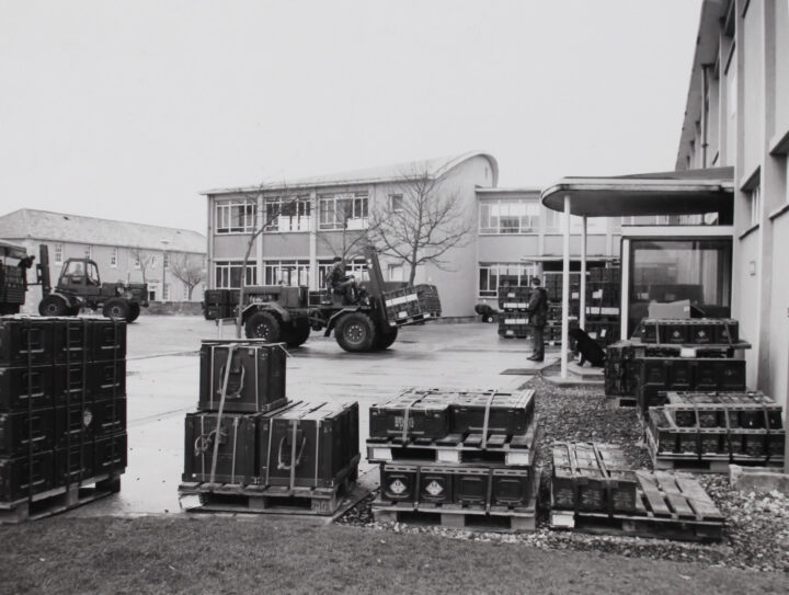 Black and white photograph of a car with a forklift listing crates in an outdoor lot. Several other crates fill the edges of the lot.