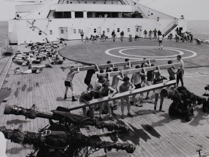 Black and white photograph of men doing athletic activities outdoors on a ship. In the foreground, groups of men lift wooden beams. In the background, a group of men are doing head and leg raises and another group are getting ready to do some running.