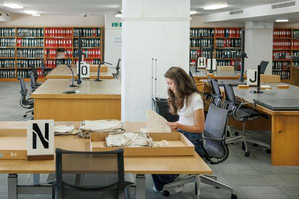A researcher sits looking at an old paper document, at a table covered with boxes.