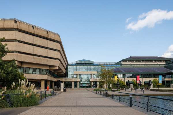 The path to The National Archives' building, with a brutalist structure on the left, a glass section in the middle, and angled roofs on the right, above a lake and some plants.