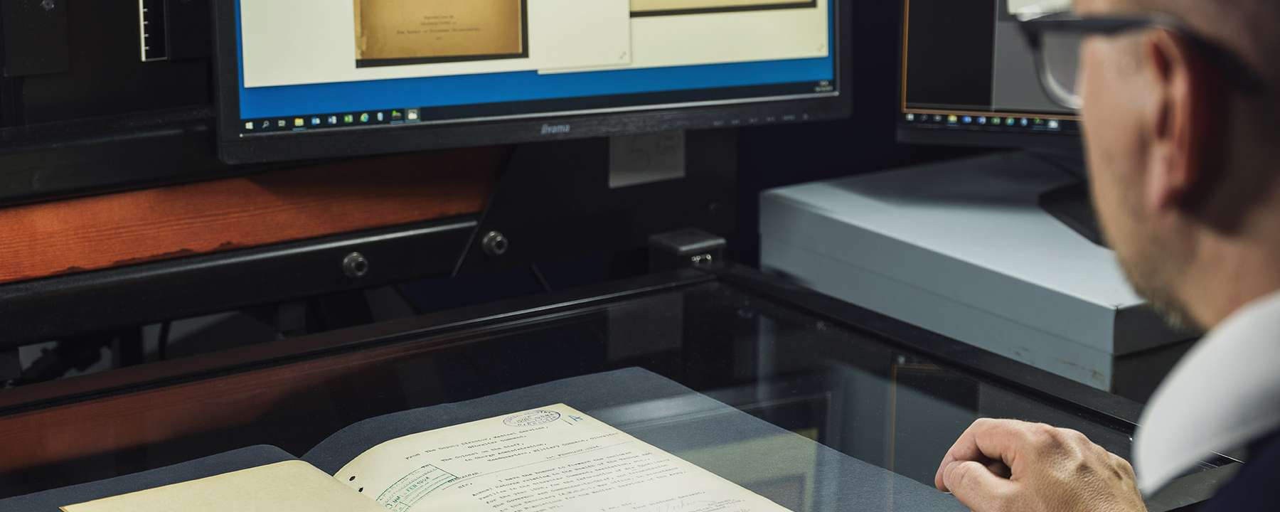 A person sits at a computer checking images of a document with the document on the table in front of them.