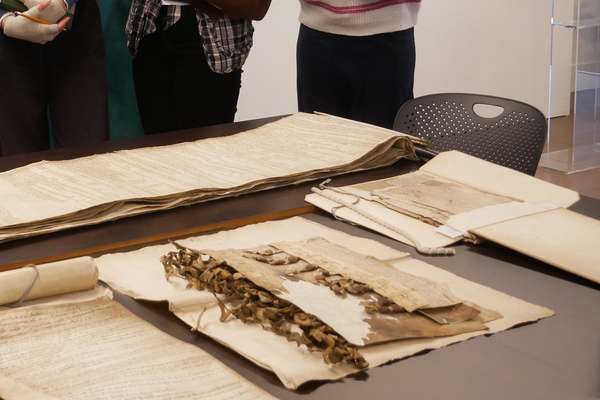 Five people standing in front of a number of records made from parchment in a educational space at The National Archives. They are inspecting the records.