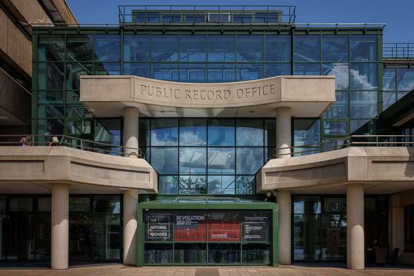 'Public Record Office' is carved in stone above the glass and metal entrance to The National Archives building, flanked by stone balconies with metal railings.