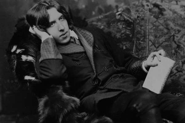 Photograph of Oscar Wilde lounging on a couch covered in fabrics and furs, holding a book in one hand.