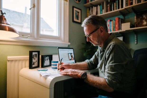 A man is sat at a desk writing on a piece of paper. He has a laptop and calculator in front of him.