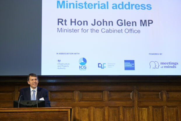 Rt. Hon. John Glen MP, Minister for the Cabinet Office, stands at a podium and speaks at the TIP Live event. Behind him, a screen shows his name and title.