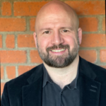 A photo of Charles Baird smiling at the camera with a brick wall behind him