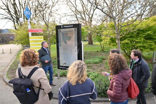 A group of 6 people – men and women – standing outdoors, looking at a large map display mounted on a stand. They appear to be looking at the map and discussing the content. Trees and a building are in the background.