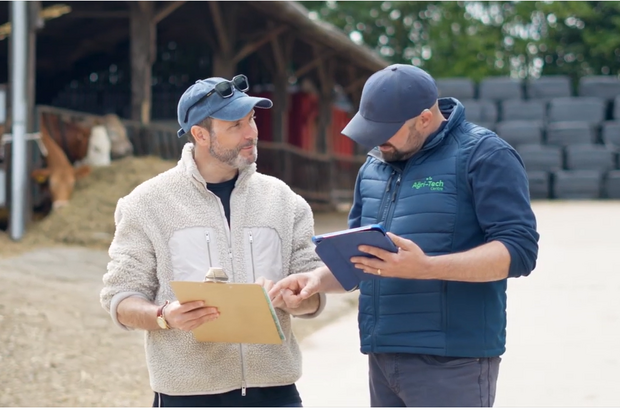 Two people standing outdoors on a farm look at documents together. One person holds a clipboard with papers, while the other points at it and holds a tablet. Behind them are a barn-like structure, stacked hay bales, and a cow partly visible in the background.