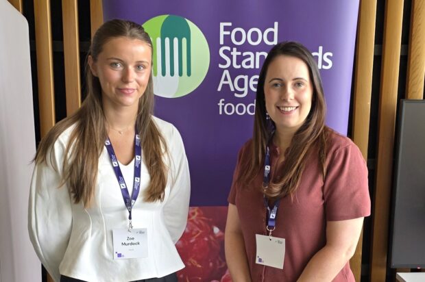 Two staff wearing lanyards stand before Food Standards Agency banner at indoor event display.