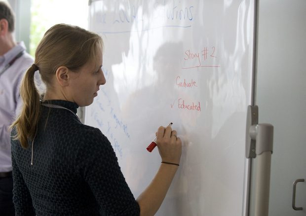 A woman writes on a whiteboard: "Story #2: Graduate, Educated (check mark)"