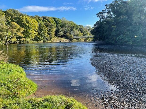 The image shows a photograph of the River Wharfe at Cromwheel, with a view to the suspension bridge across the river.
