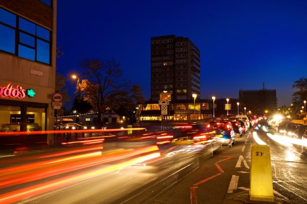 Cars driving down a busy main road in a town centre, in the dark with their lights on.