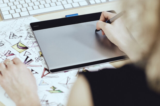 Photo of a hand using a pen on a graphics tablet to draw an unseen illustration. Tablet is set on a desk in front of a keyboard and surrounded by sheets of paper featuring a range of hand-drawn illustration examples.