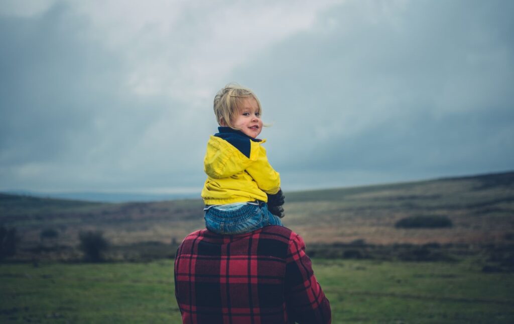 A toddler in a yellow mac sits on an adult's shoulders, against the backdrop of a cloudy sky