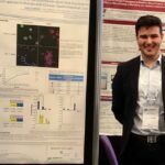 Image of a male in a suit standing in from of posters containing text, images of bacteria and graphs.
