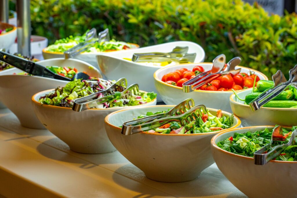 A buffet-style salad bar with large white serving bowls filled with fresh vegetables, including mixed leafy greens, sliced tomatoes, cucumbers and chopped salad ingredients. Metal serving tongs rest in each bowl.
