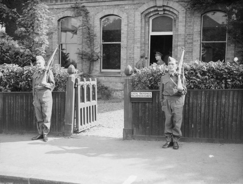 Two members of the Local Defence Volunteer Corps, bearing rifles and standing guard outside a building. An officer can be seen talking with another one of the ranks near the entrance.