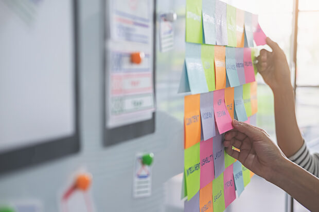 A whiteboard full of colourful post-it notes with hands placing new notes onto the surface