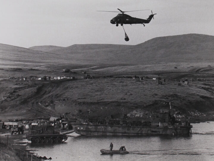 Black and white photograph of a helicopter flying with cargo hanging beneath it overall coastal area. There is a ship and tank in the water next to the shore, and trucks on the shore. Tire tracks lead to a small settlement further inland.
