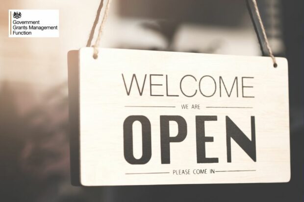 A shop sign reading Welcome we are open. Please come in - with the Government Grants Management Function logo in the top left corner