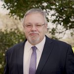 Image of Chris Hadkiss. He is dressed in a navy suit, white shirt and purple tie and is standing against a background of trees.