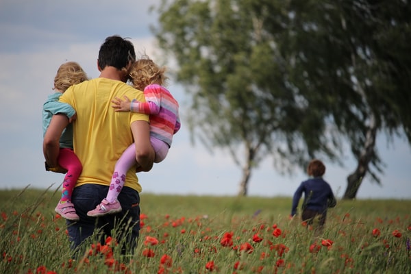 Father walking through field, carrying two children, with one child running ahead. Photo by Juliane Liebermann on Unsplash