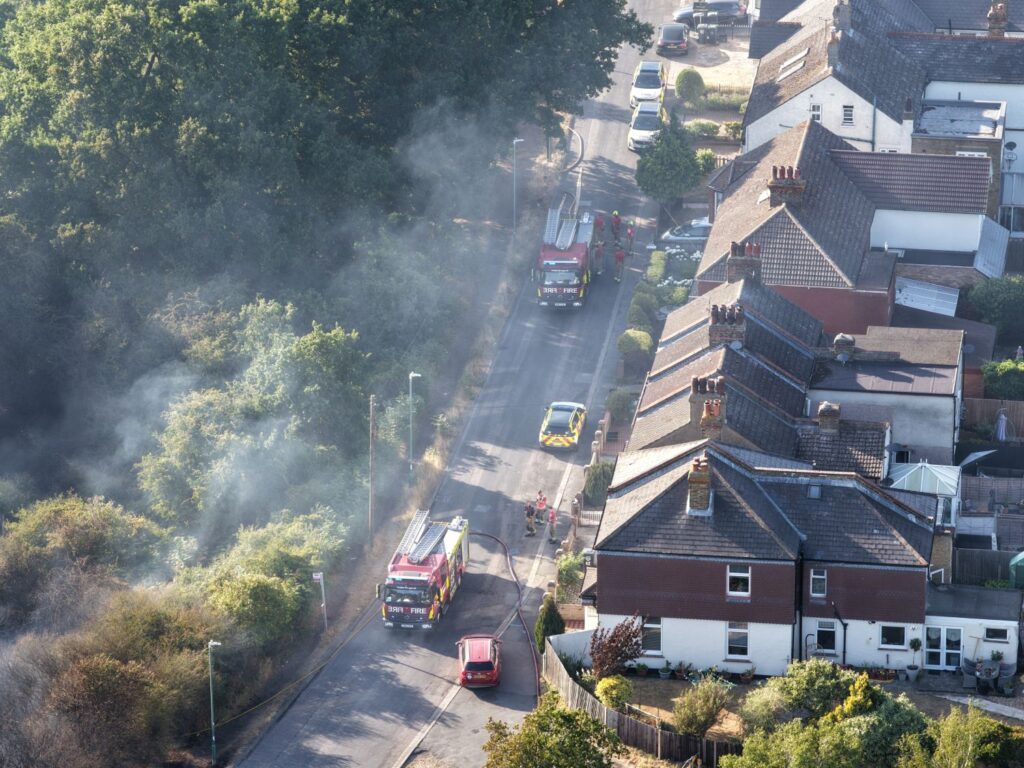 Aerial view of a residential street bordered by dense vegetation, with two fire engines and an emergency vehicle responding to a wildfire in Kent. Smoke drifts from the vegetation beside the road, while rows of houses line the opposite side and firefighters work near the vehicles.