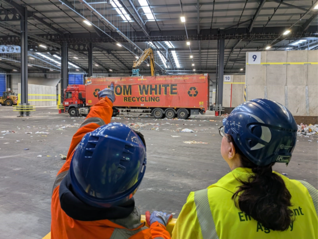 Chief Regulator Jo Nettleton alongside a site manager - both wearing hard hats and hi-vis jackets - at an indoor waste management facility. The site manager is pointing to AI-sensors on the ceiling that are used to support the sorting of waste.