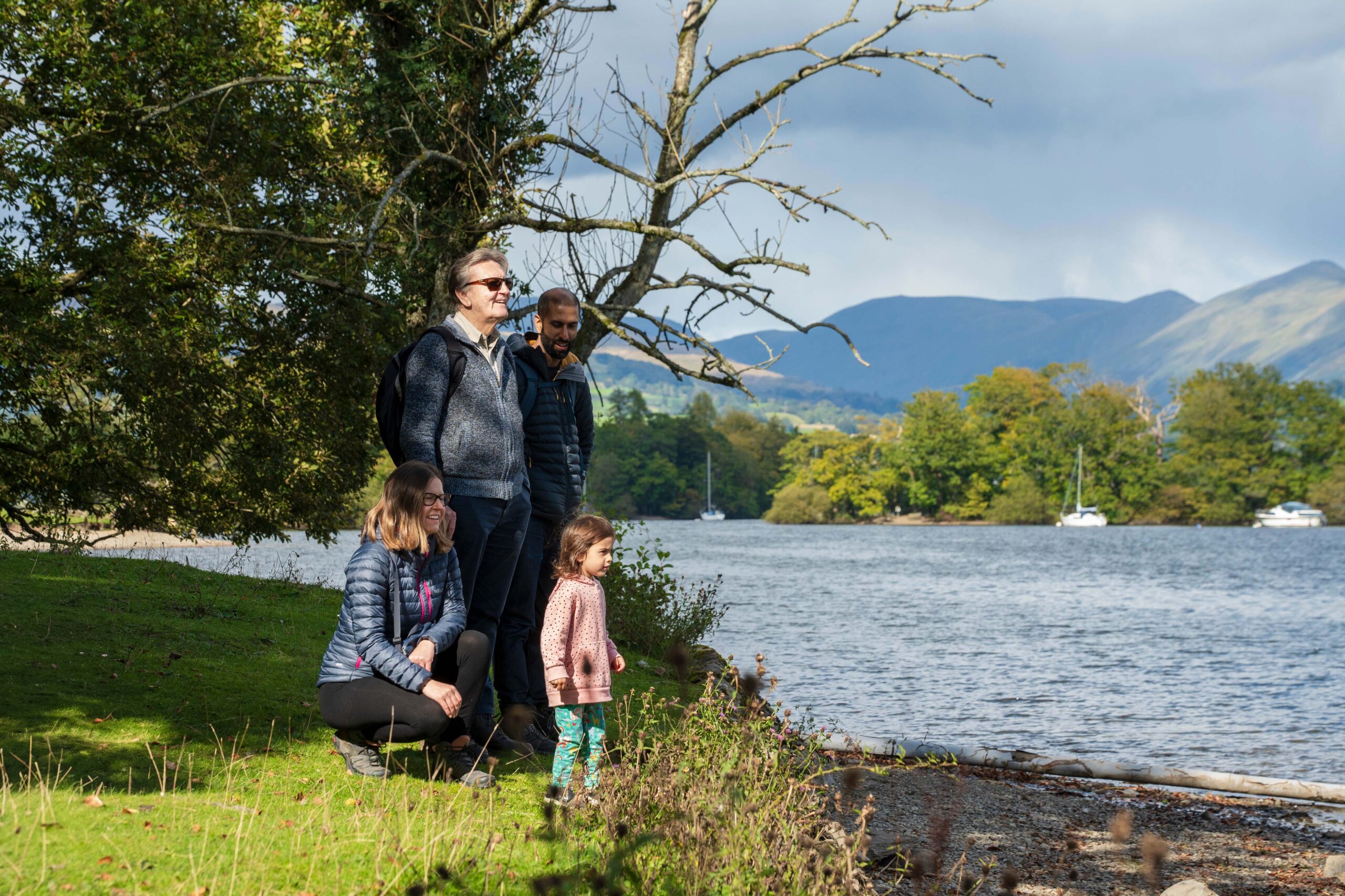 child, adult female, adult male, elderly male, stood together on grass lake front under green tree, looking out over a lake on a sunny day