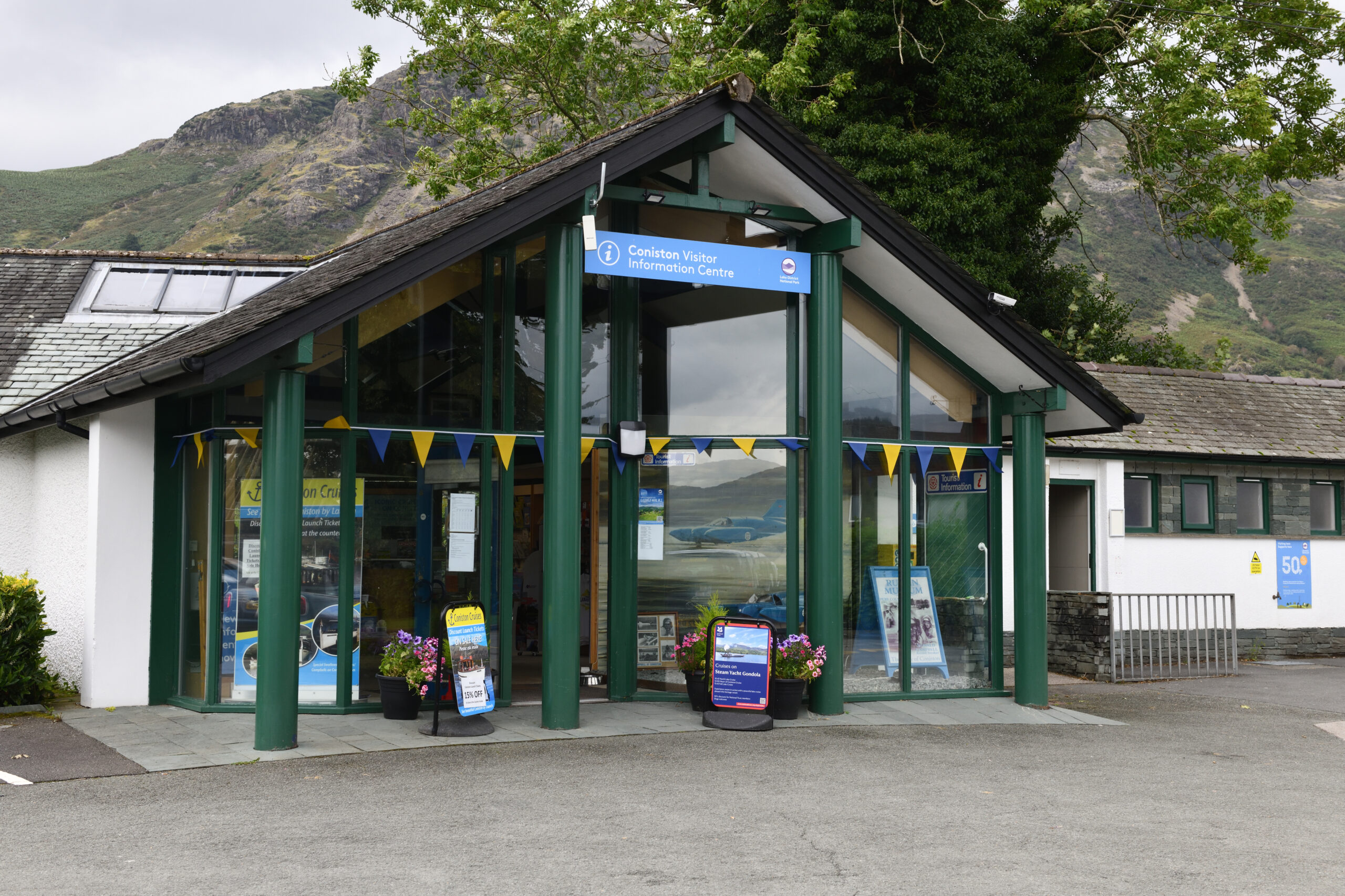 Outside the front of Coniston Information Centre. Triangular roof framing large green wooden framed glass windows. Blue centre sign at the top of the door. Yellow and navy triangle bunting strung mid-way across the entrance. Grey tarmac ground surrounding