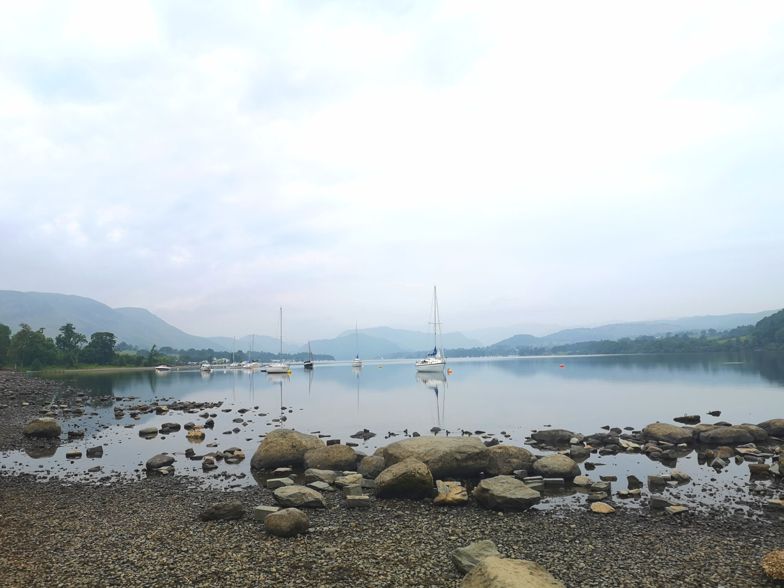 Pebbled shore of Ullswater near Pooley Bridge looking over the still lake with sailing boats in the distance