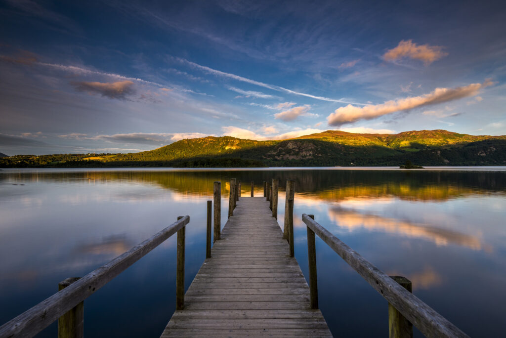 Looking down a wooden jetty into Derwent Water. The lake is blue and orange, reflecting the sunset sky and woodland hills above