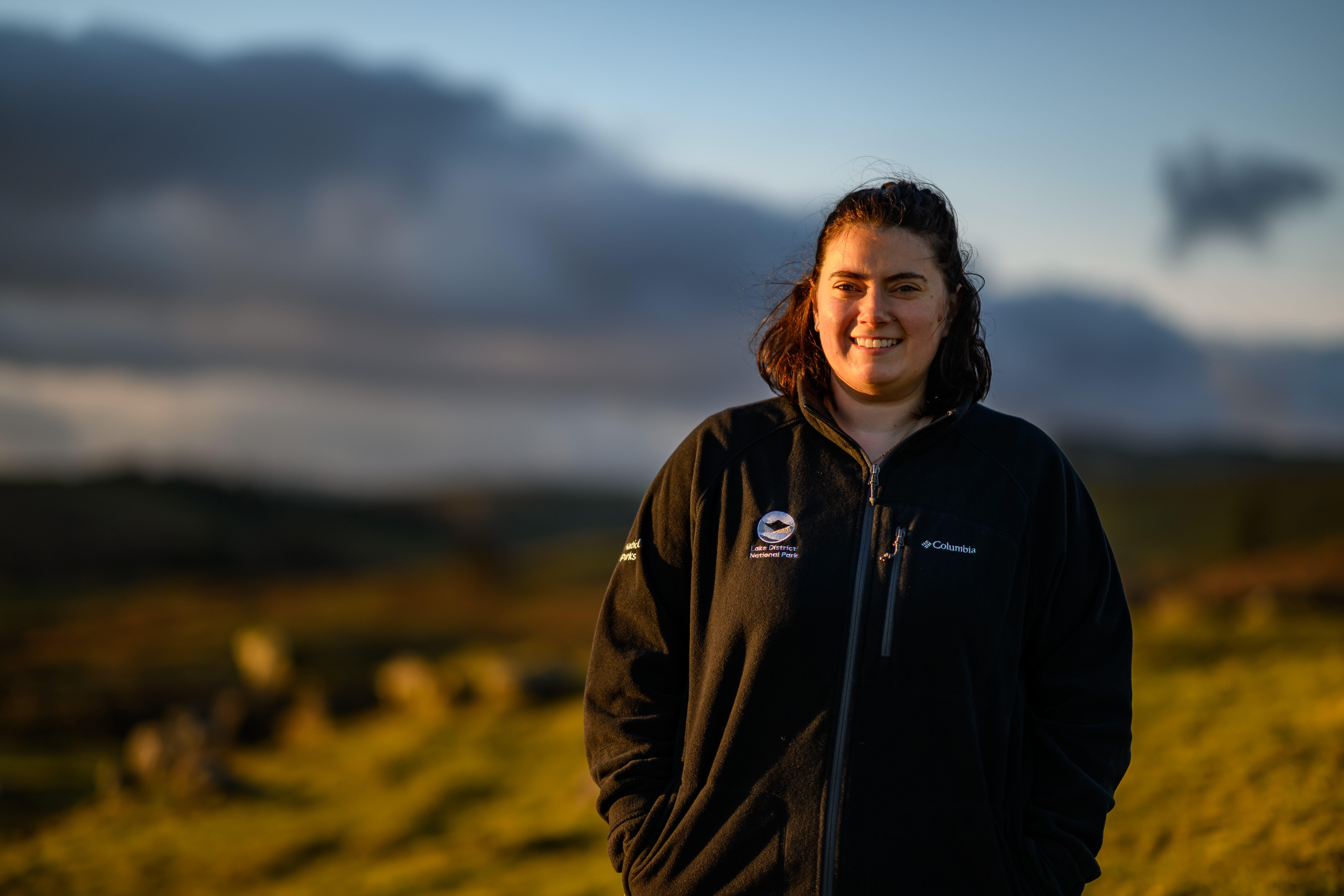 adult female in branded black fleece smiling stood on grassy fell