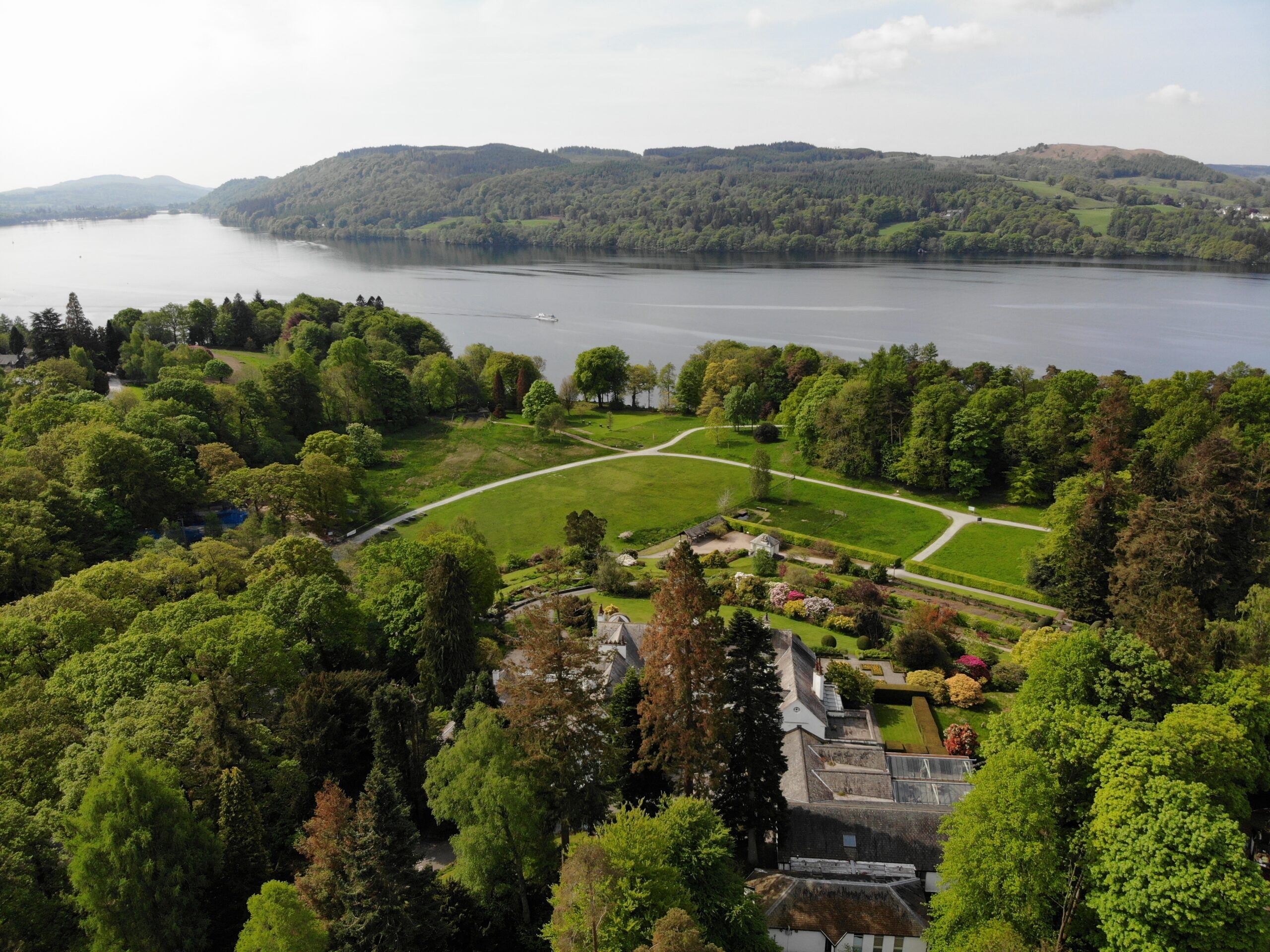aerial view of brockhole house and gardens. lush green grass and woodland with intersecting stone paths. windermere lake behind