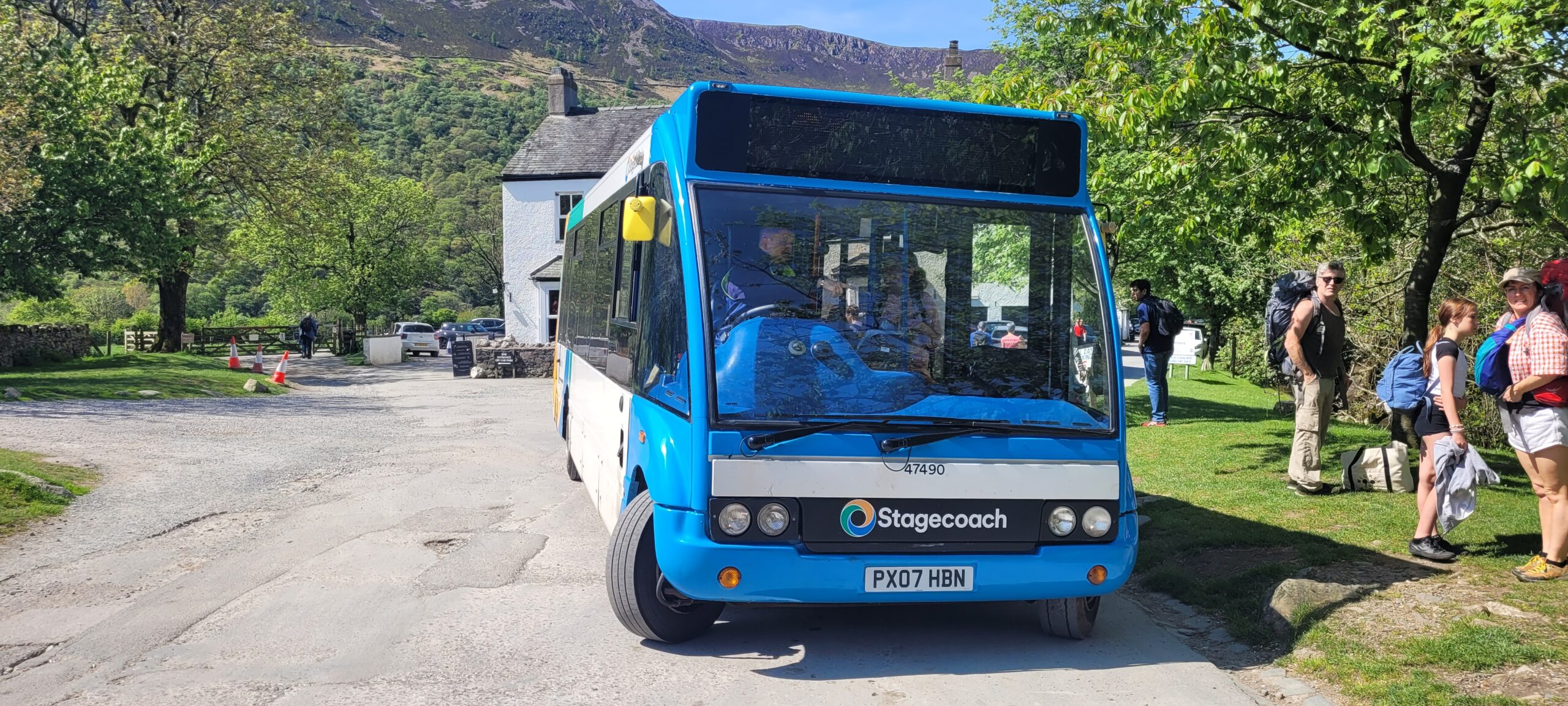 blue buttermere shuttlebus parked on concrete with passengers diembarked on grass to right, white stone house and fells behind bus