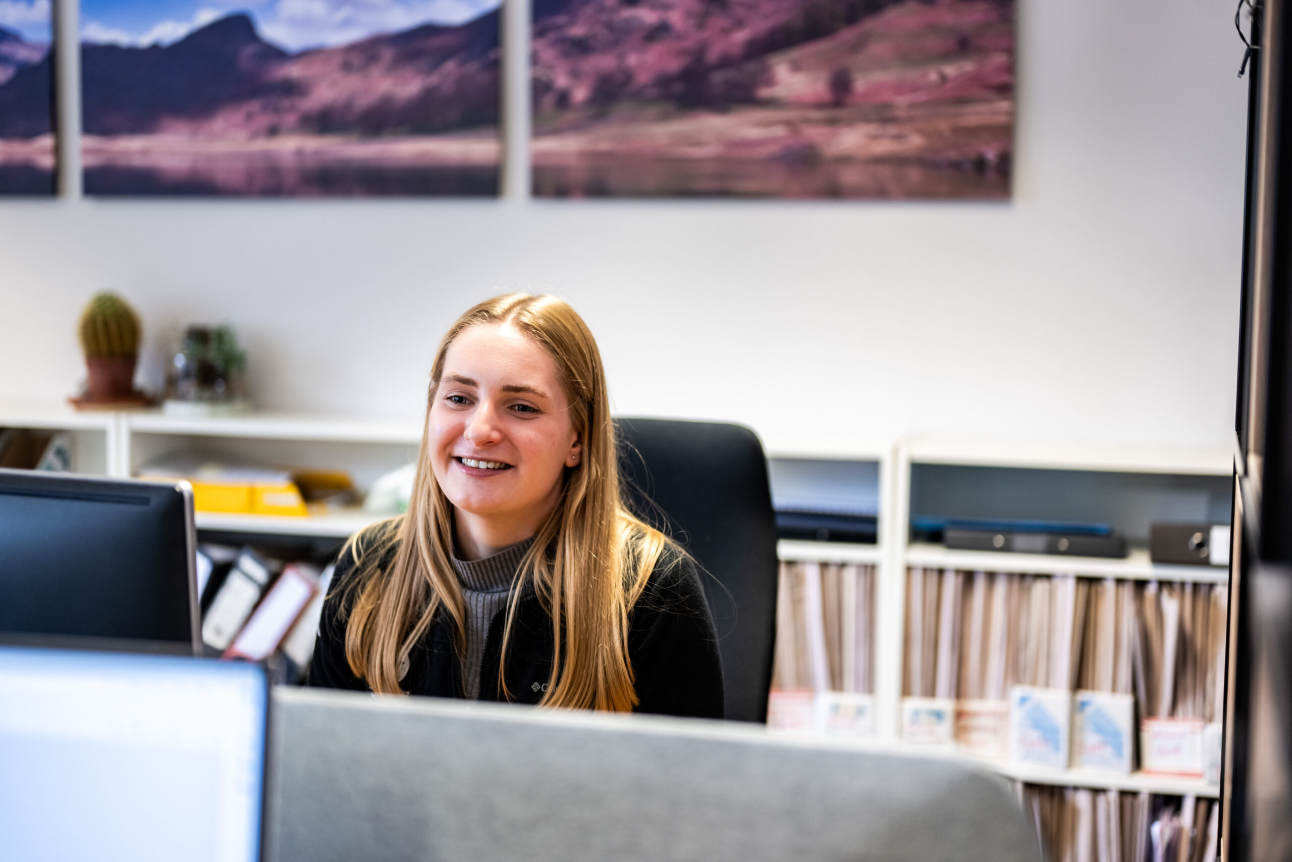 A blond woman sat at a desk looking at a computer