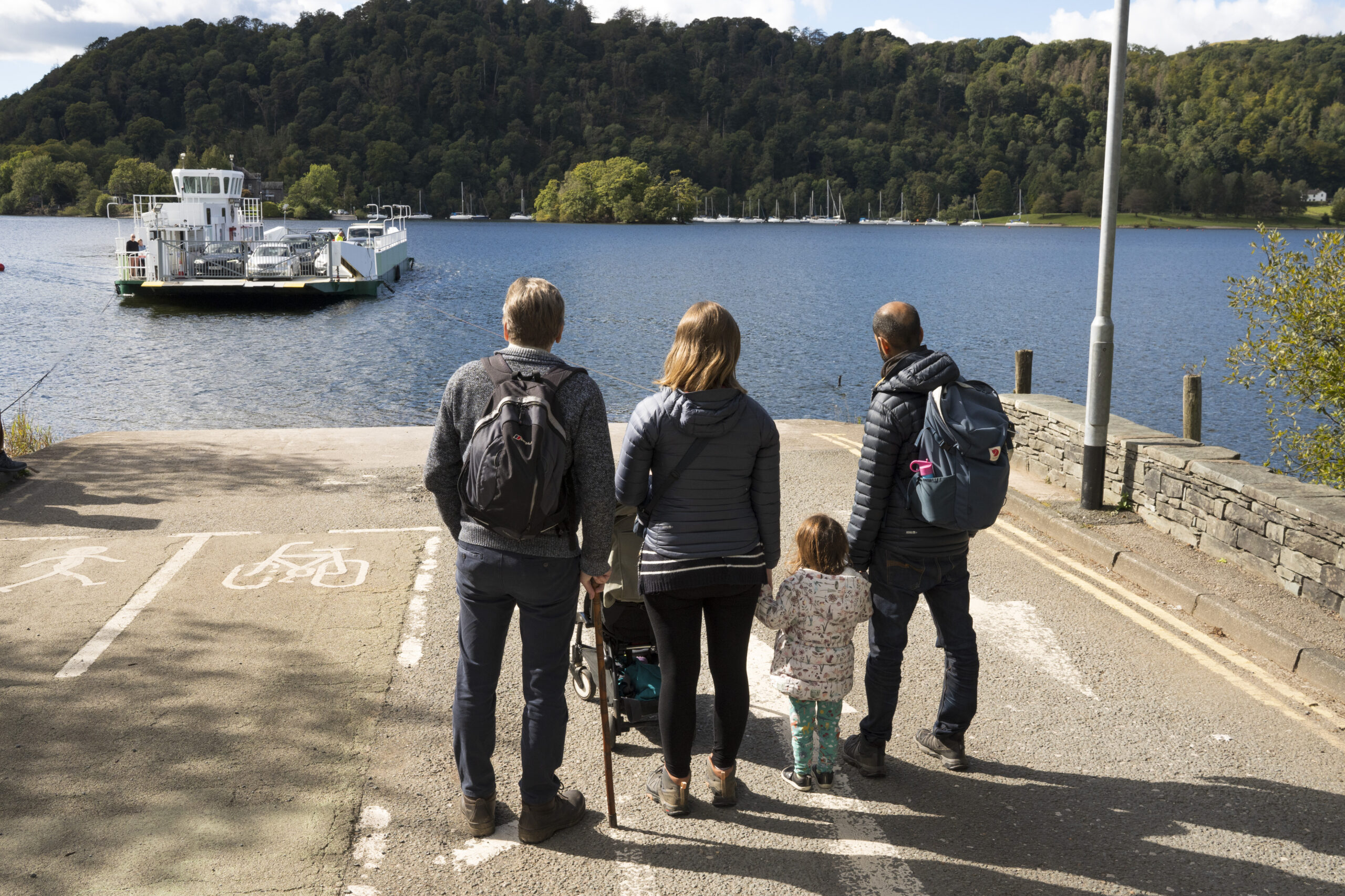 three adults and child waiting for the ferry in a passenger waiting area. looking out to windermere lake, surrounded by green trees
