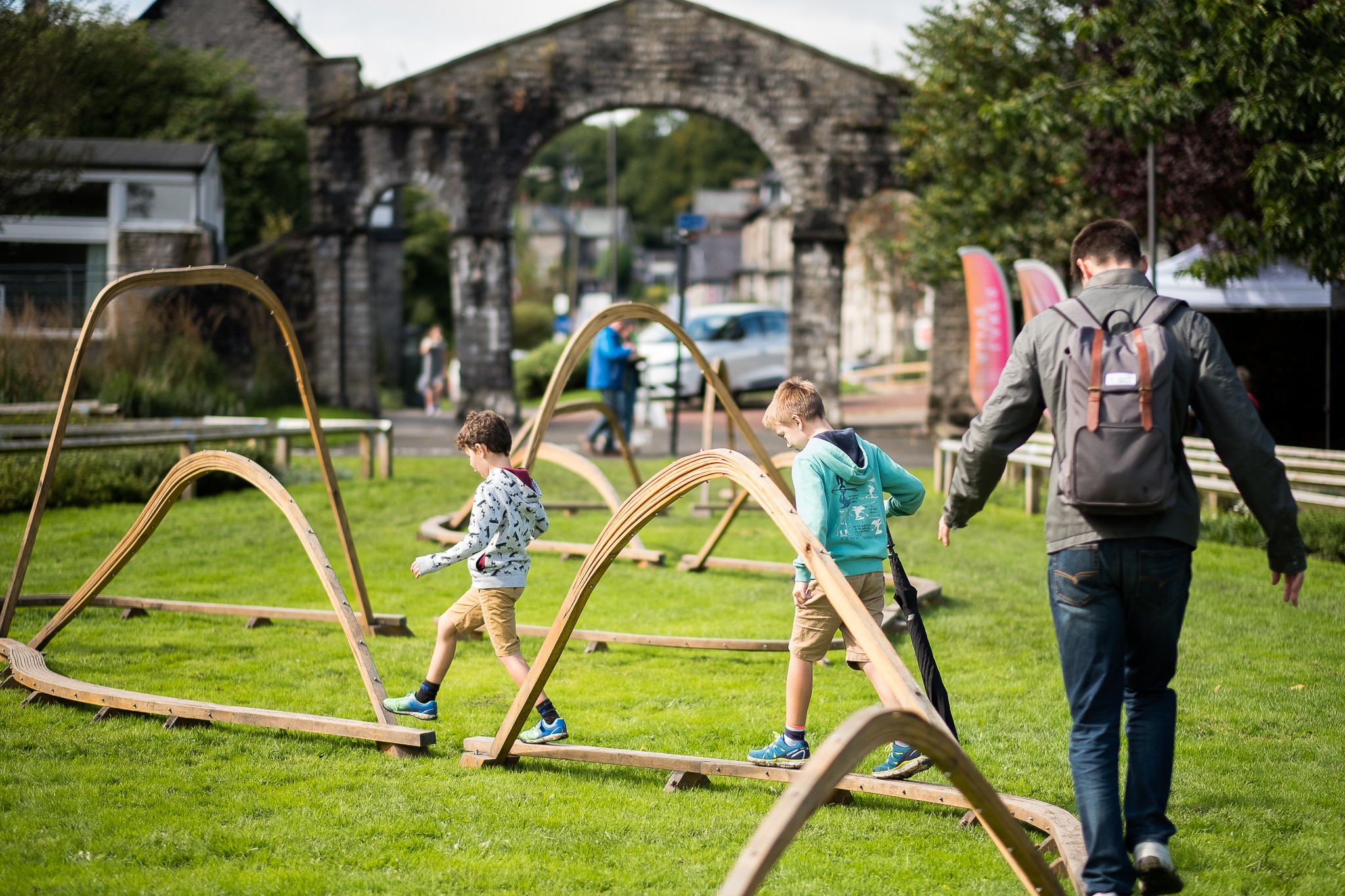 Adult and two children playing at the Lakes Alive event. The children are balancing on wooden structures, in a green field, in front of a stone arch and gate