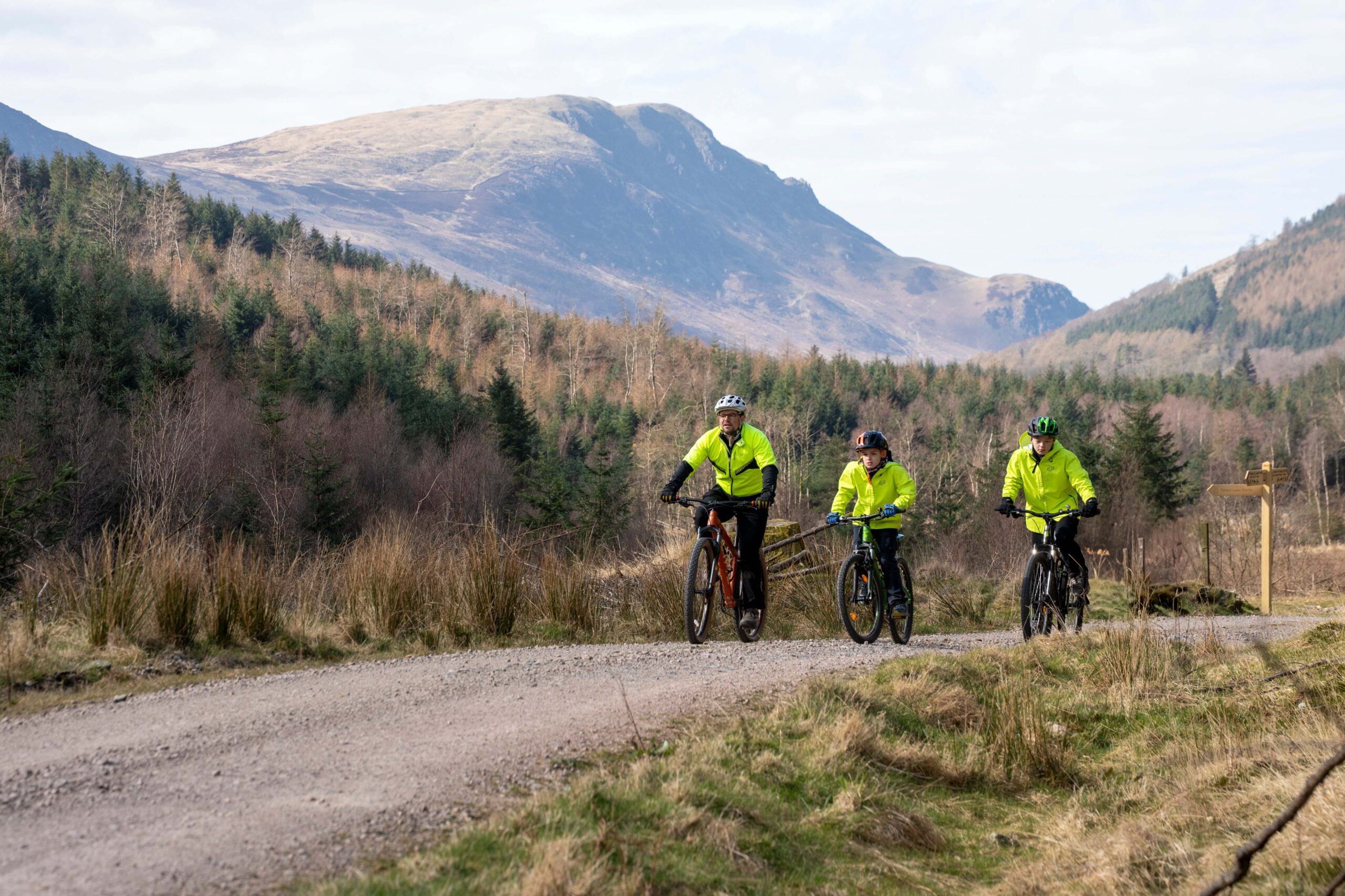 adults male and two boys in yellow high-vis cycling on wide gravel track with fell backdrop