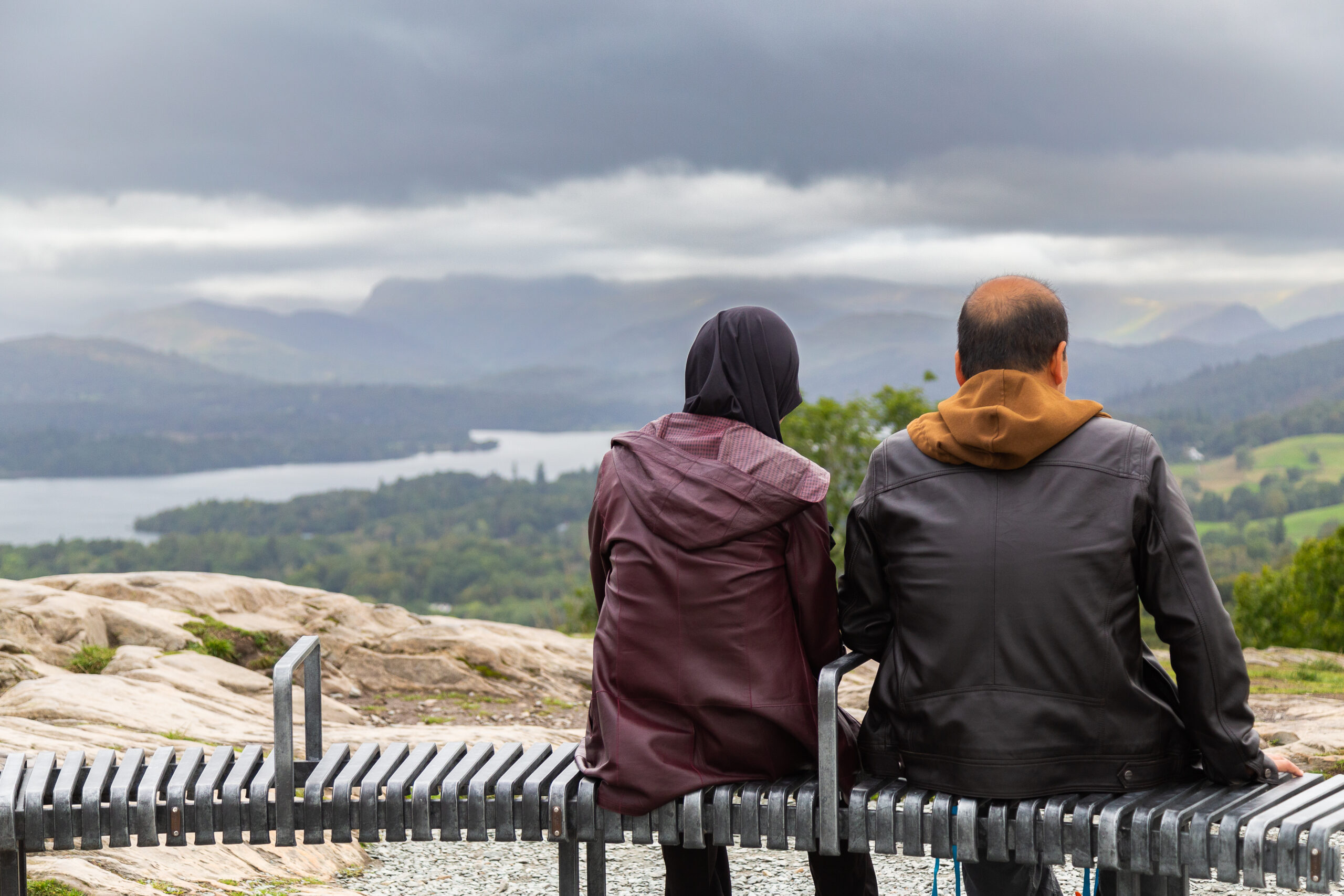 A couple sat on the bench at the top of Orrest Head looking across to the Lakeland fells over Windermere