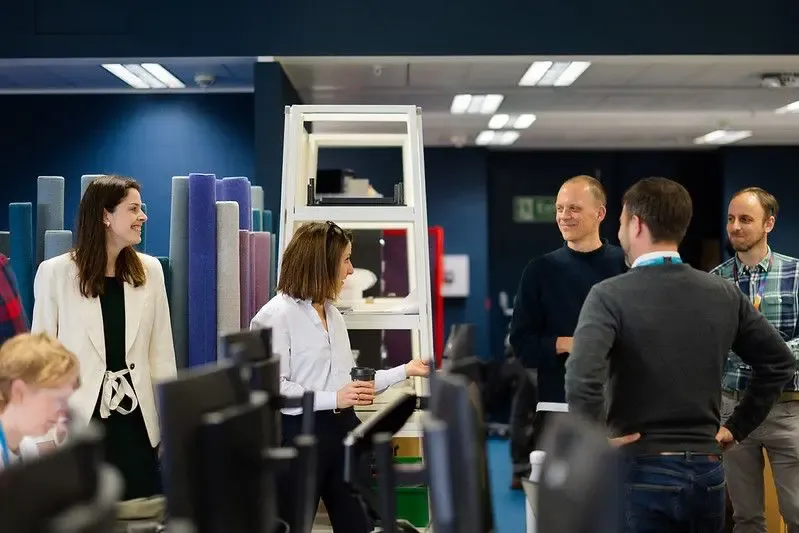 Members of the i.AI team in an office standing up and smiling as one member talks to the rest
