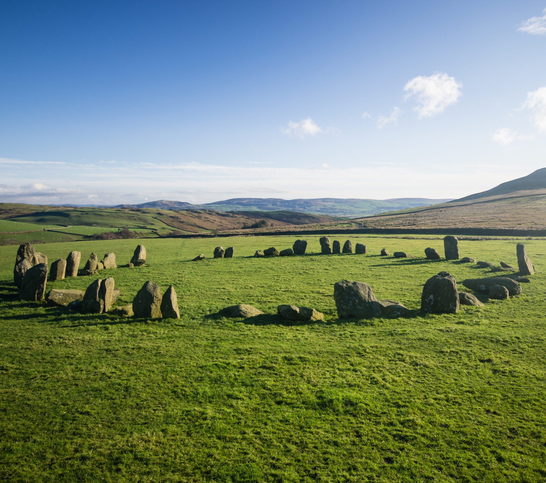 A stone circle in the middle of a green field on a bright sunny day, with a blue sky and a handful of clouds