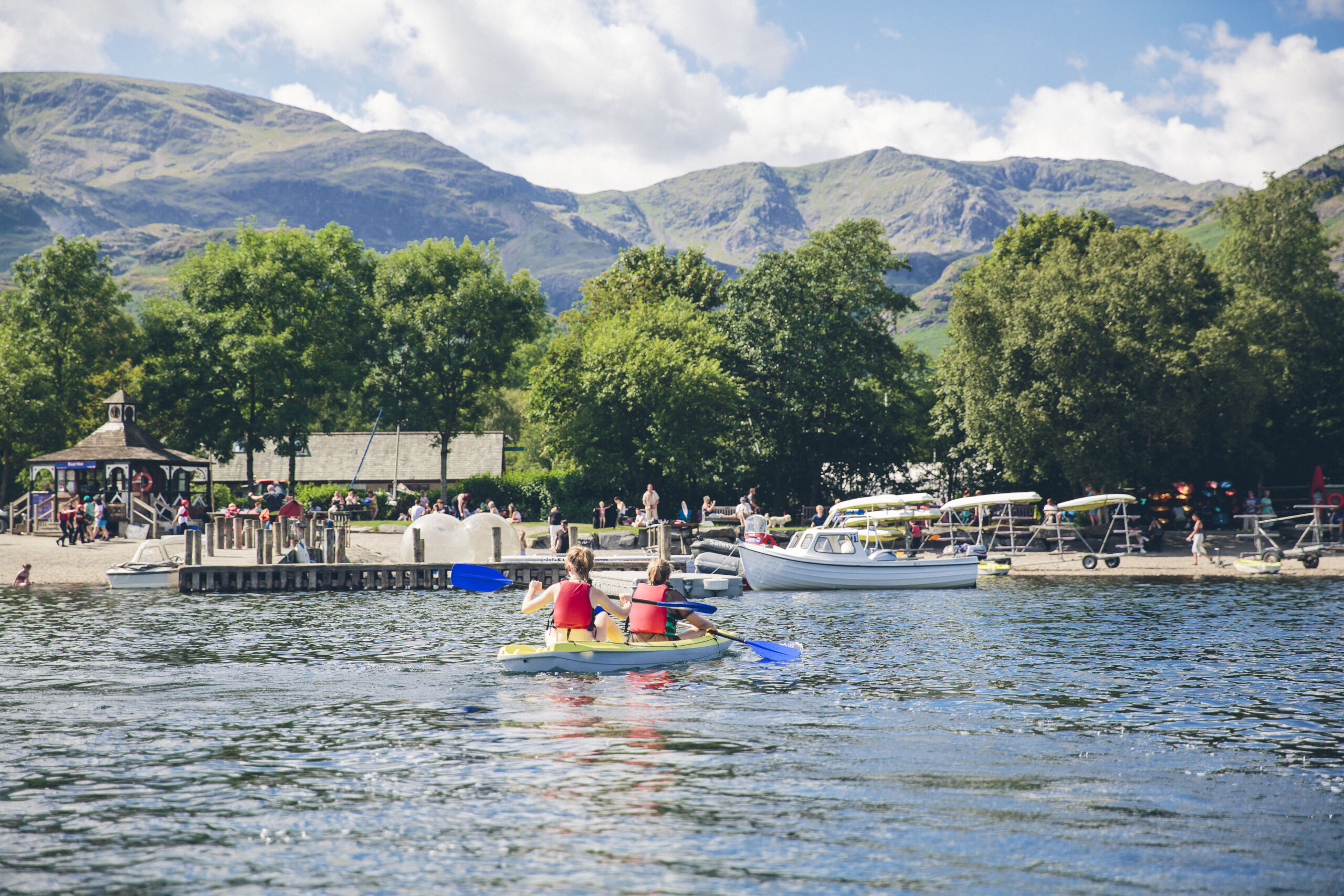 View of Coniston Boating Centre. Sunny day. On the blue water is two older children in red buoyancy aids kayaking, a moored white motorised boat and a wooden jetty. On the lake shore is are canoe and kayak transportation trolleys, two shelter huts and many people. Behind are green trees and green fells.