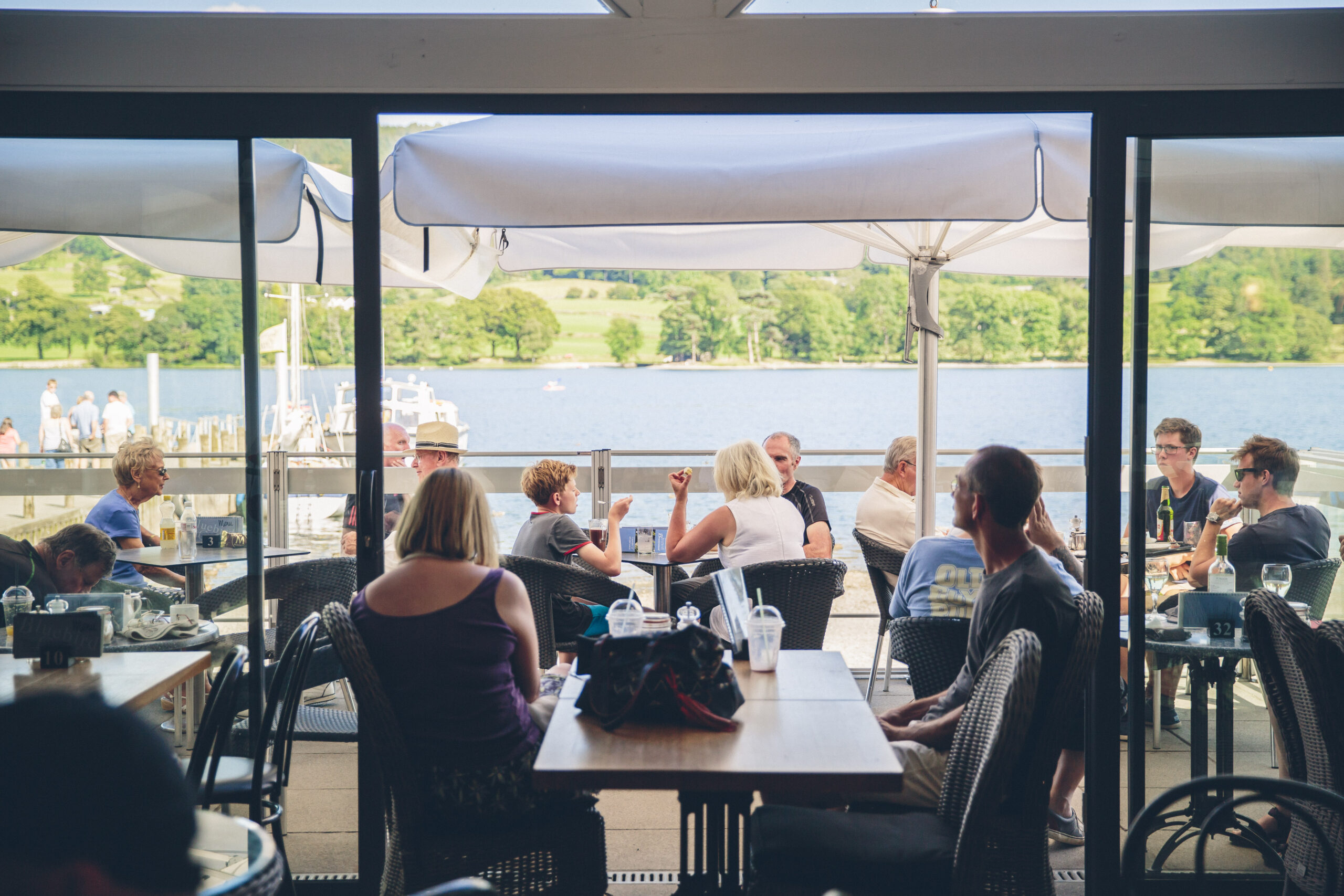 Inside The Bluebird Café. Many adults and families sat at tables, looking out of and around the large glass floor-to-ceiling windows, overlooking a sunny Coniston Water