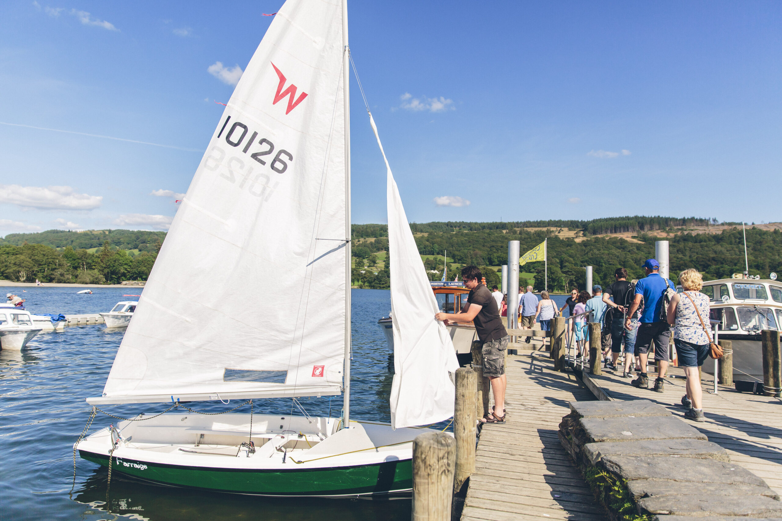 Green and white sailing boat with large white sail moored next to wooden jetty on Coniston Water. Adult male attends to sail as visitors walk down the jetty. Sunny blue sky with green fells in the background
