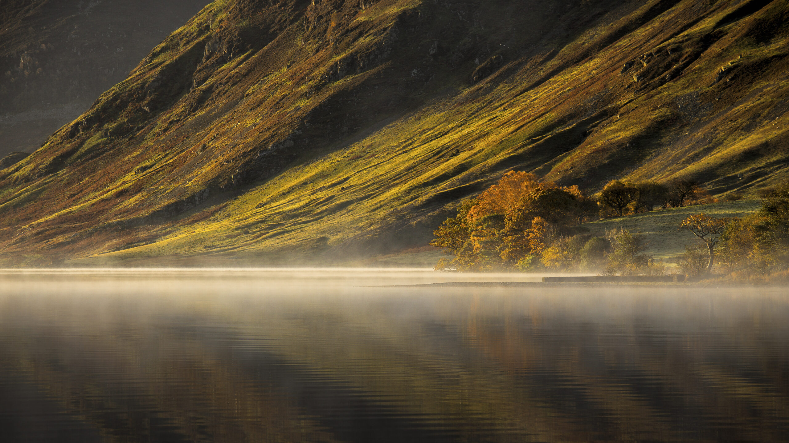 orange lit fell slope reflecting into crummock water, mist and autumnal woodland lines the lake shore