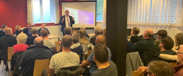 A man speaking at the front of a room to a large group of people.