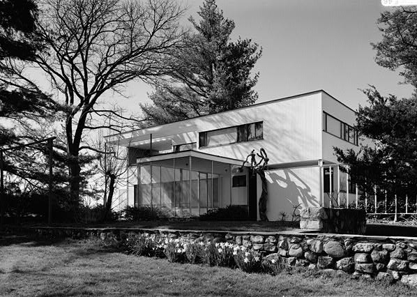 A black-and-white photograph of the Walter Gropius House, from 1938, located at 68 Baker Bridge Road in Lincoln, Massachusetts. The house, designed by architect Walter Gropius, is a modernist structure with a flat roof, clean lines, and rectangular forms. It features large windows and a partially glass-walled entrance area, emphasising natural light and connection to the outdoors. A minimalist balcony with an open-frame structure extends above the entryway, while a stone retaining wall borders a landscaped garden in the foreground. The surrounding trees and shrubs frame the house, blending the architecture with its natural setting.