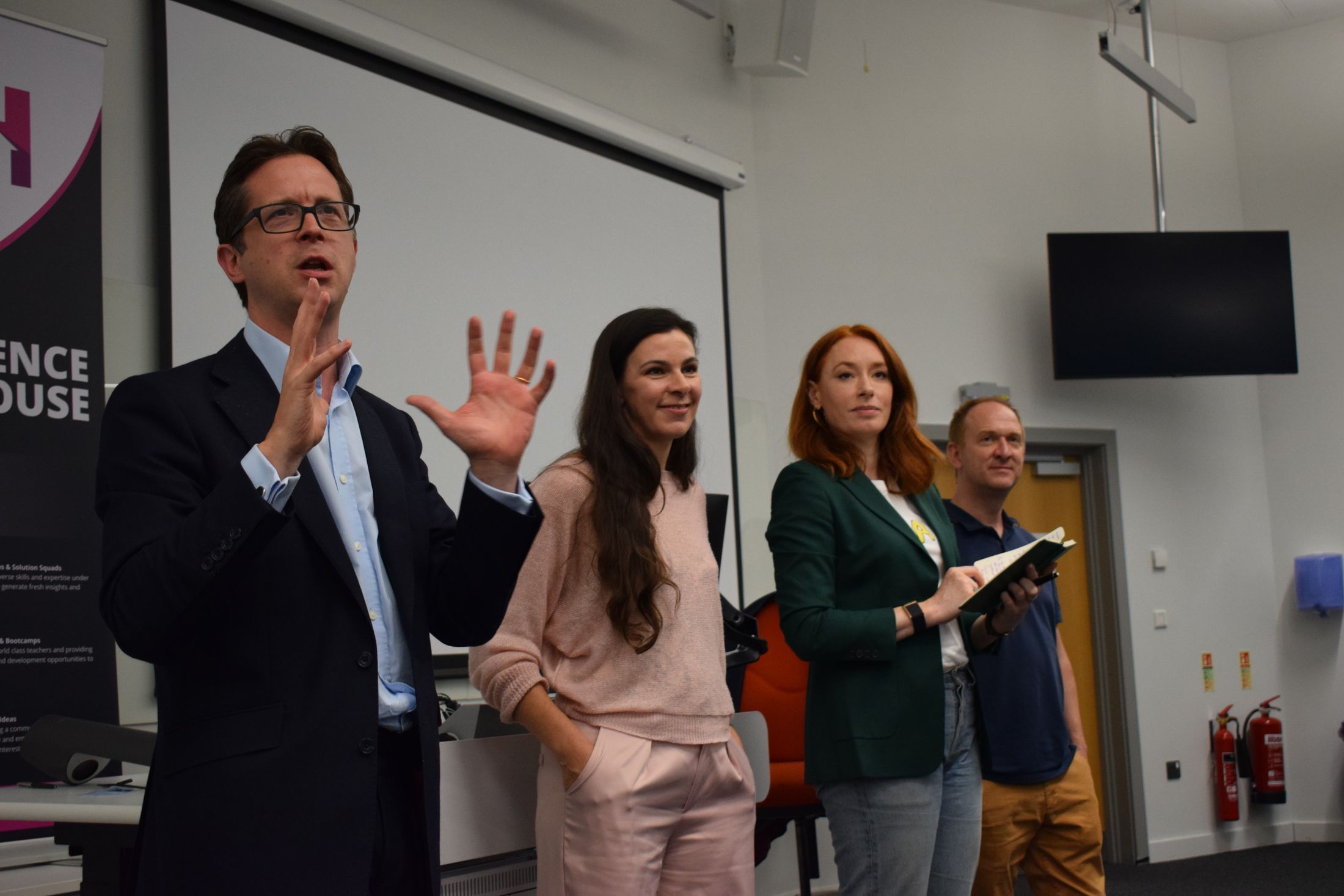 Alex Burghart, Laura Gilbert, Hannah Fry and Tom Read stood up at the front of the room. Alex Burghart is speaking
